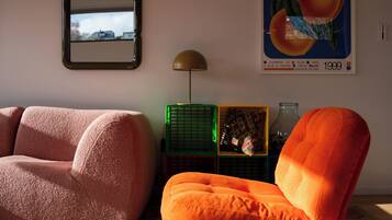 Ein Wohnzimmer mit einer Couch, einem orangefarbenen Sessel, einem Spiegel, einer Lampe, einem Poster und einem gemusterten Teppich.
