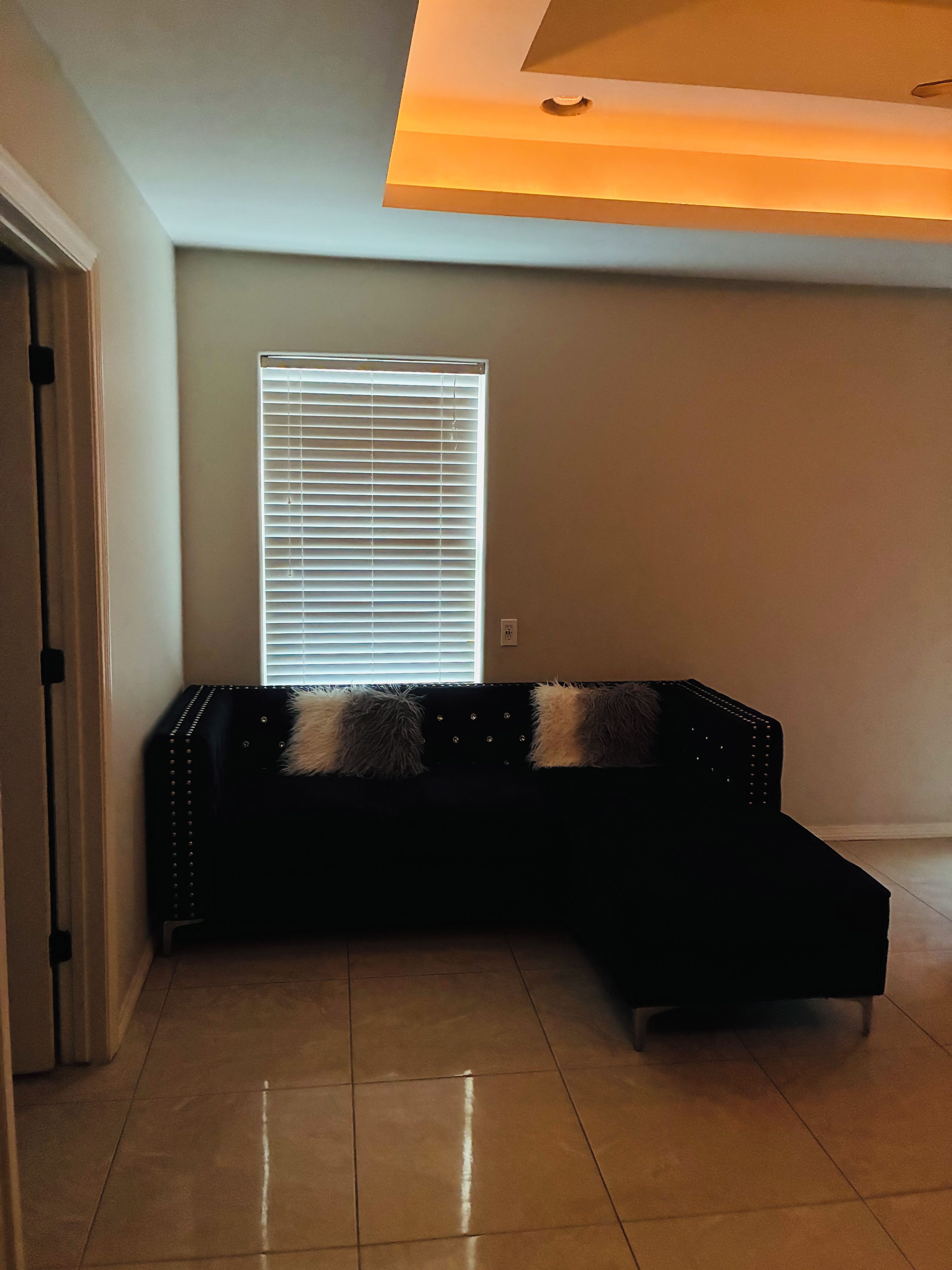 Ein Zimmer mit einer schwarzen Polstersofa, einem Fenster mit Jalousien und einem Fliesenboden.