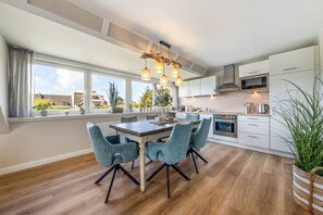 A modern kitchen with a dining area, large windows, and a view of the neighborhood. - Sylter Herzstück - Modern 3-room vacation apartment with approximately 75 m² of living space, for (Wenningstedt-Braderup (Sylt))