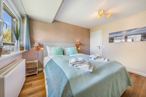Ein Schlafzimmer mit Bett, Nachttischlampen, einem Heizkörper, einem Fenster mit Vorhängen und einem Wandbild.