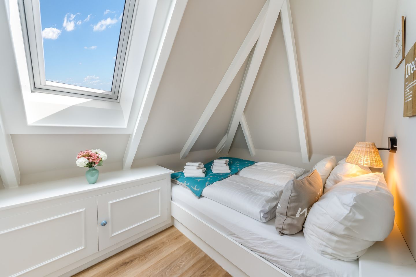 Un dormitorio con techo inclinado, una cama con ropa blanca, una mesita de noche, una lámpara y un ventanal con vistas al cielo.
