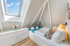 A bedroom with a sloped ceiling, a bed with white bedding, a nightstand, a lamp, and a window with a view of the sky. - Sylter Herzstück - Modern 3-room vacation apartment with approximately 75 m² of living space, for (Wenningstedt-Braderup (Sylt))