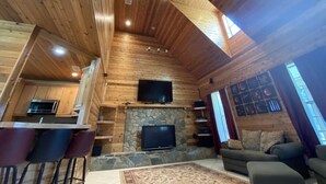 A cabin interior with a stone fireplace, a television mounted above it, a dining area with bar stools, and a sofa in the living room. - Spacious 4BR Ocala National Forest Home with Private Hot Tub Near Salt Springs — Sleeps 8 (Salt Springs)