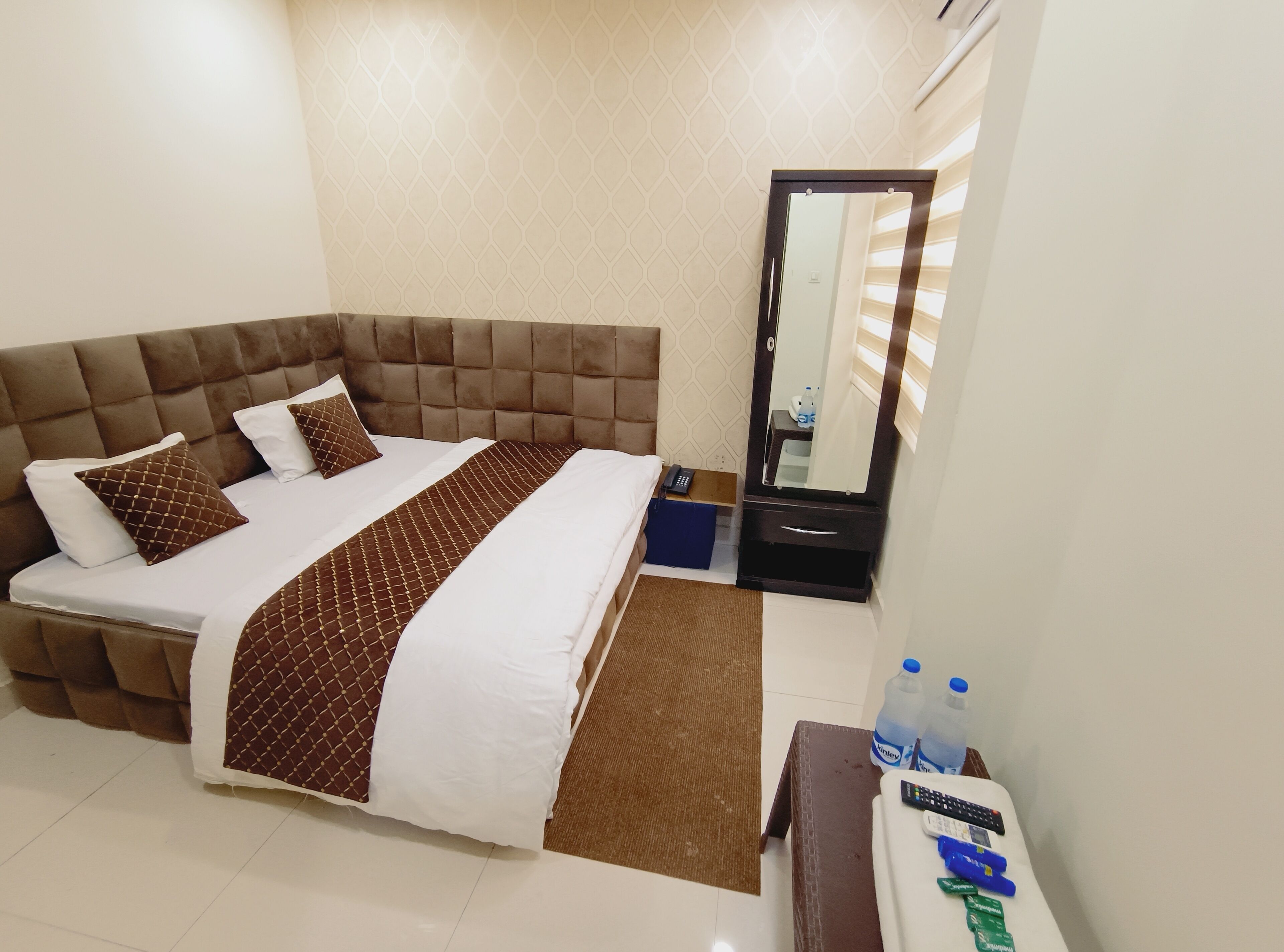 A hotel room with a large bed, a bedside table with a mirror, and a small table with a remote control and water bottles.