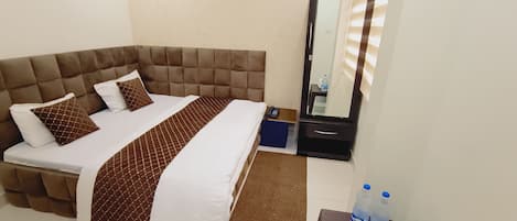 A hotel room with a large bed, a bedside table with a mirror, and a small table with a remote control and water bottles.