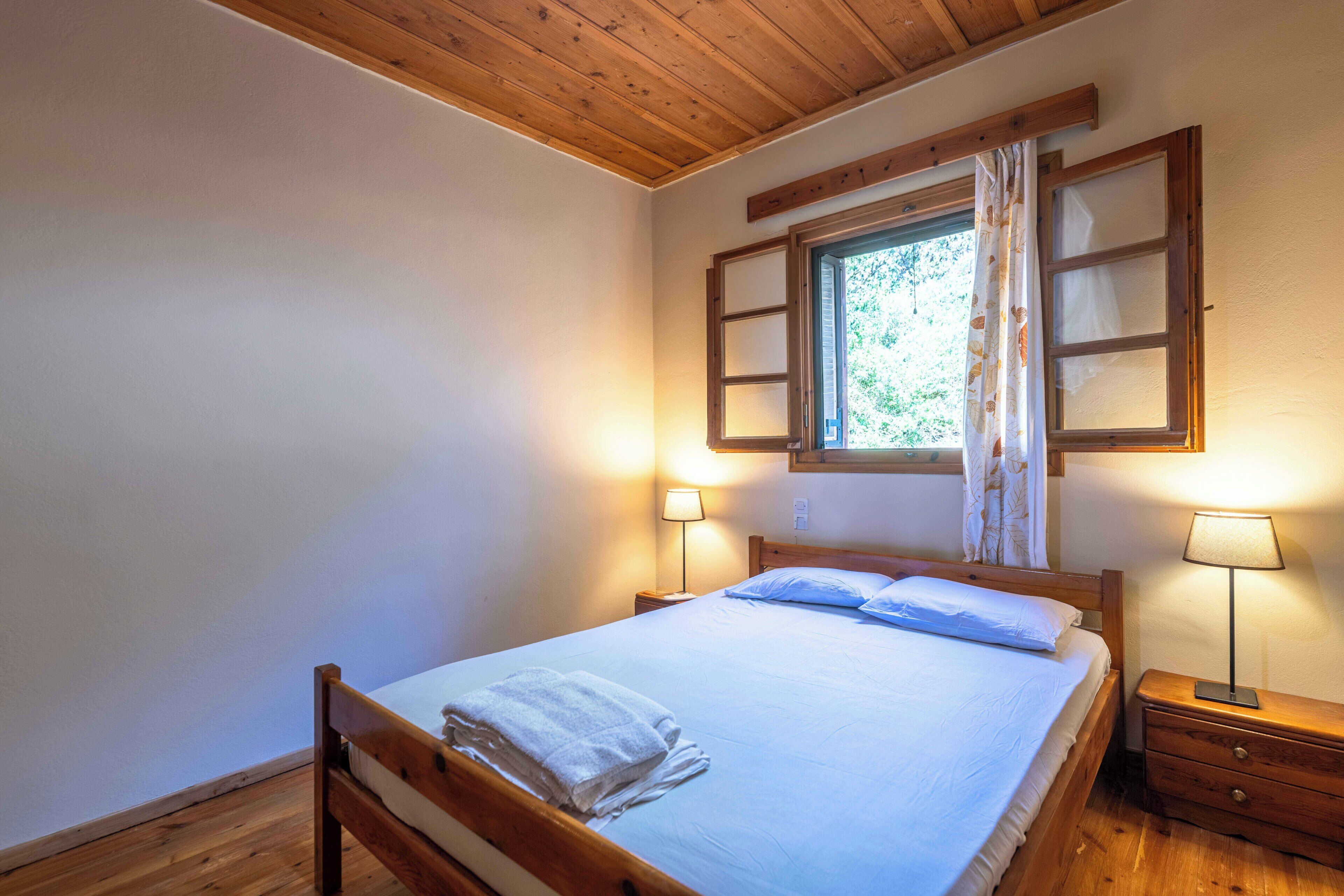 Uma cama de solteiro com cabeceira de madeira, duas luminárias de cabeceira, uma janela com venezianas abertas e uma mesa de cabeceira de madeira.