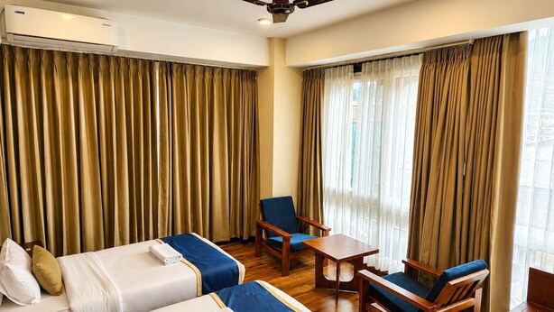 Una habitación de hotel con dos camas, un ventilador de techo, una mesita pequeña, una silla y una ventana con cortinas.