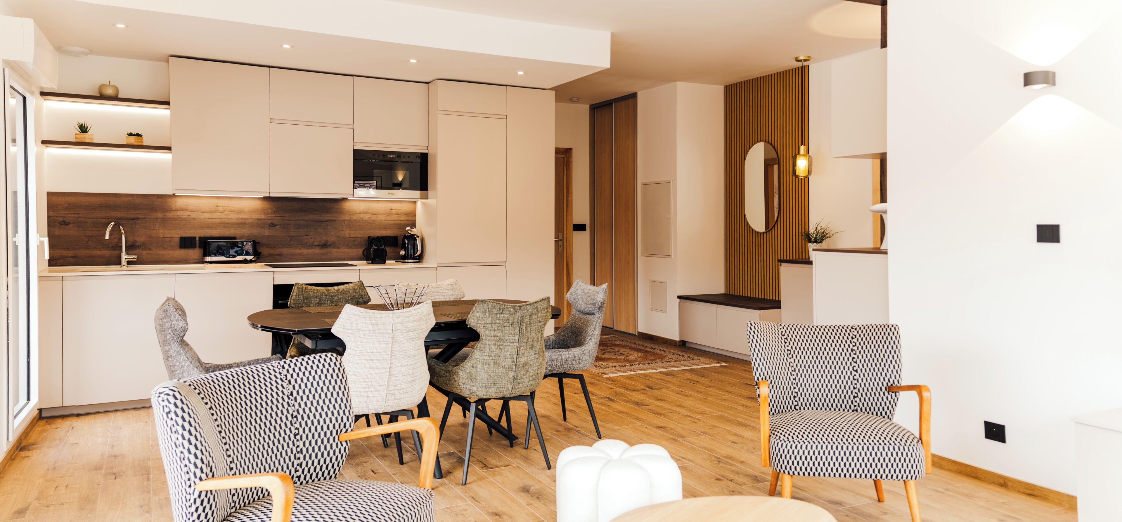 A modern kitchen with built-in appliances, a dining area with patterned chairs, and a wooden floor.