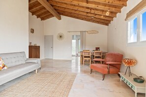 A spacious living area with a high wooden ceiling, a grey sofa, a red armchair, a dining table with chairs, and a floor lamp.