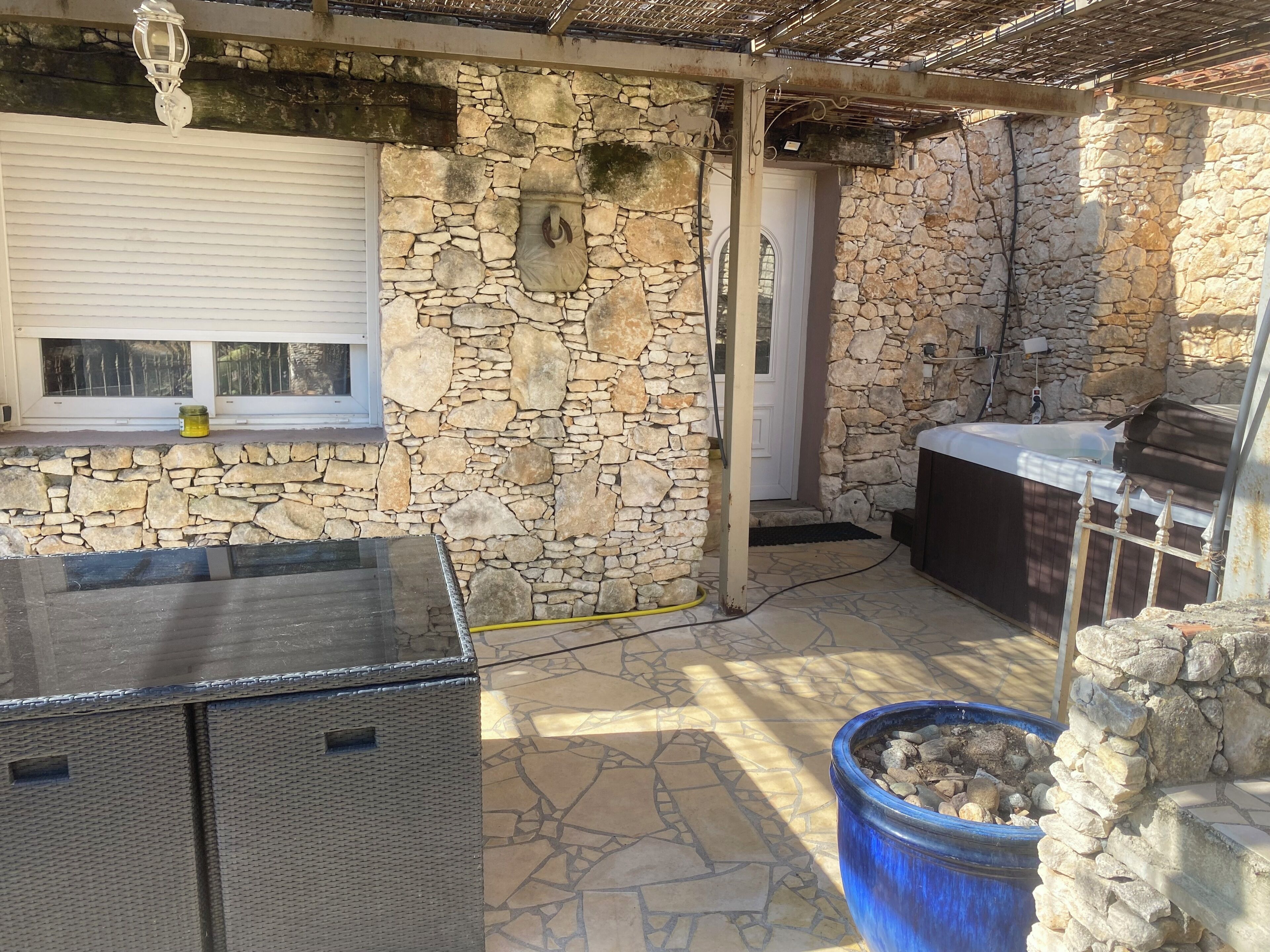 A stone-walled outdoor area with a hot tub, a wicker table, and a large blue pot.