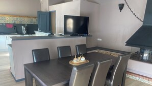 Uma área de refeições com mesa, cadeiras, televisão e uma cozinha ao fundo.