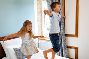 Two children jumping on a bed in a room with a window, curtains, and a radiator.