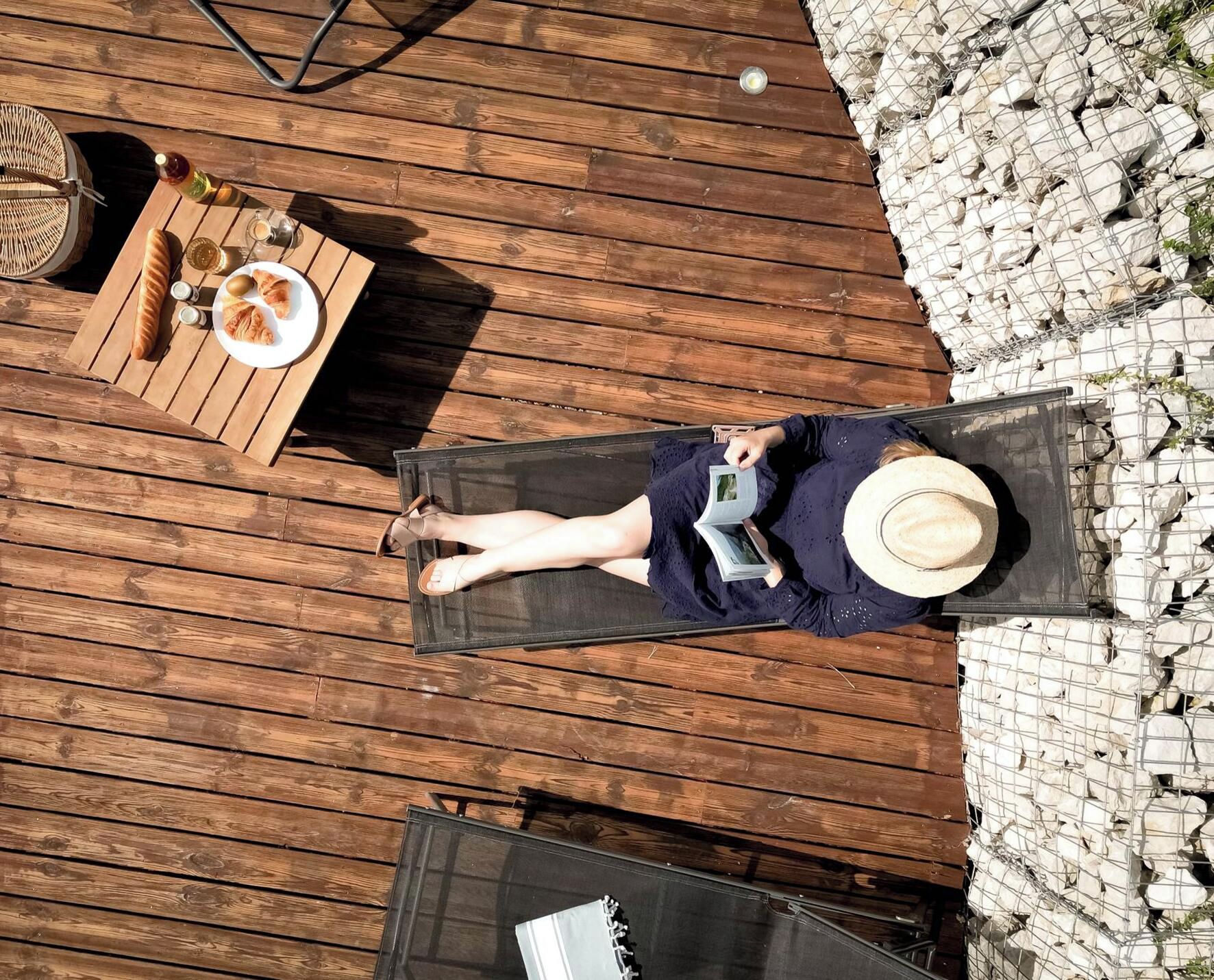 Una persona recostada sobre una cubierta de madera, con sombrero, leyendo un libro.