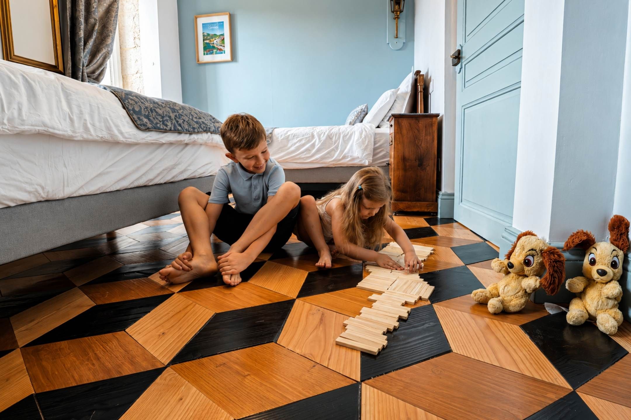 Deux enfants jouent avec des blocs de bois sur un sol à carreaux, dans une chambre où se trouvent un lit, une table de chevet et une lampe fixée au mur.