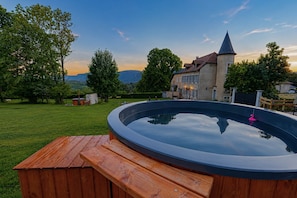 Un jacuzzi con un muelle de madera, rodeado por un jardín césped y árboles, con un edificio y montañas al fondo.