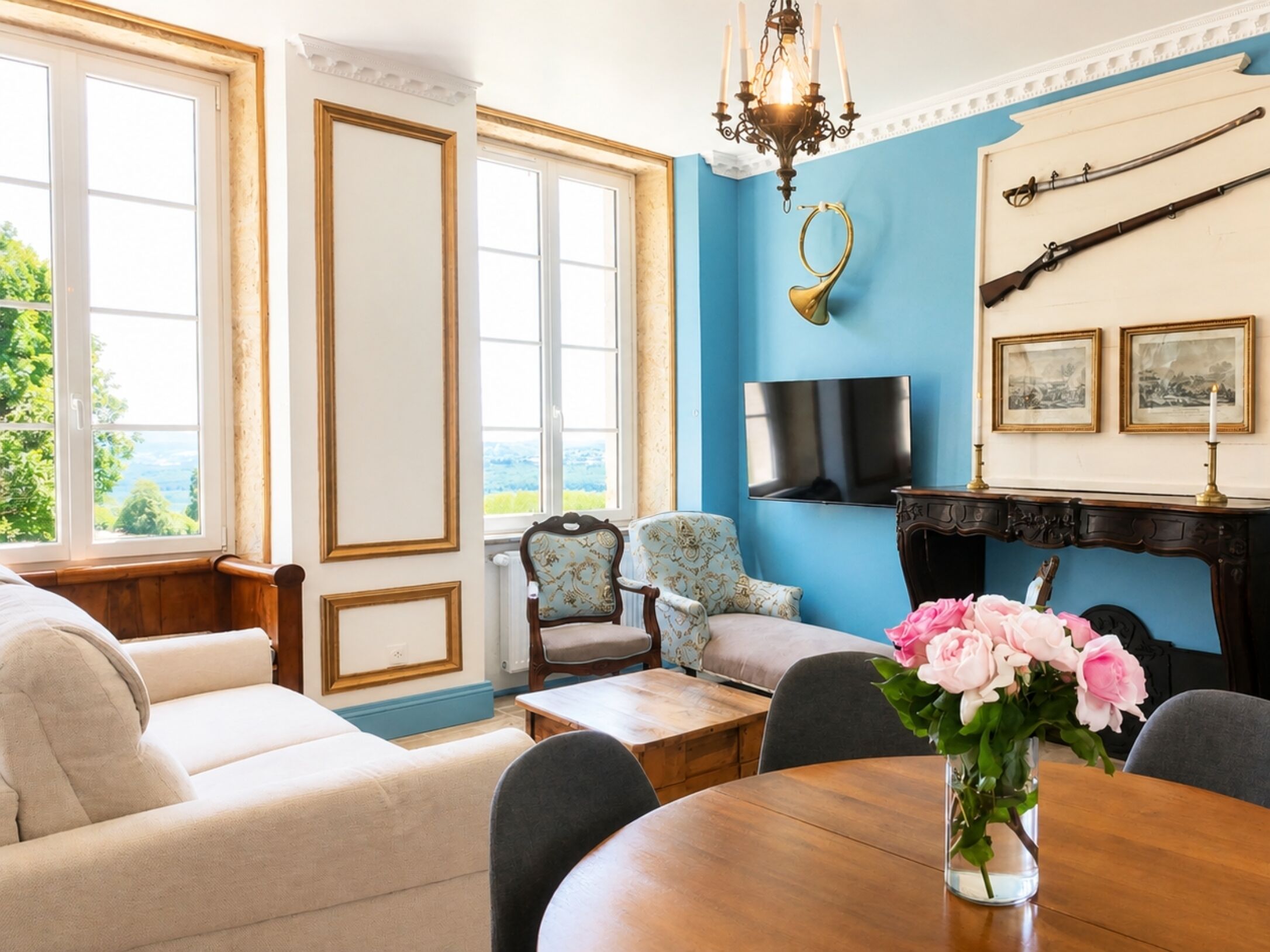 A room with a blue accent wall, a fireplace with mounted weapons, a television, and a large window with a view of greenery.