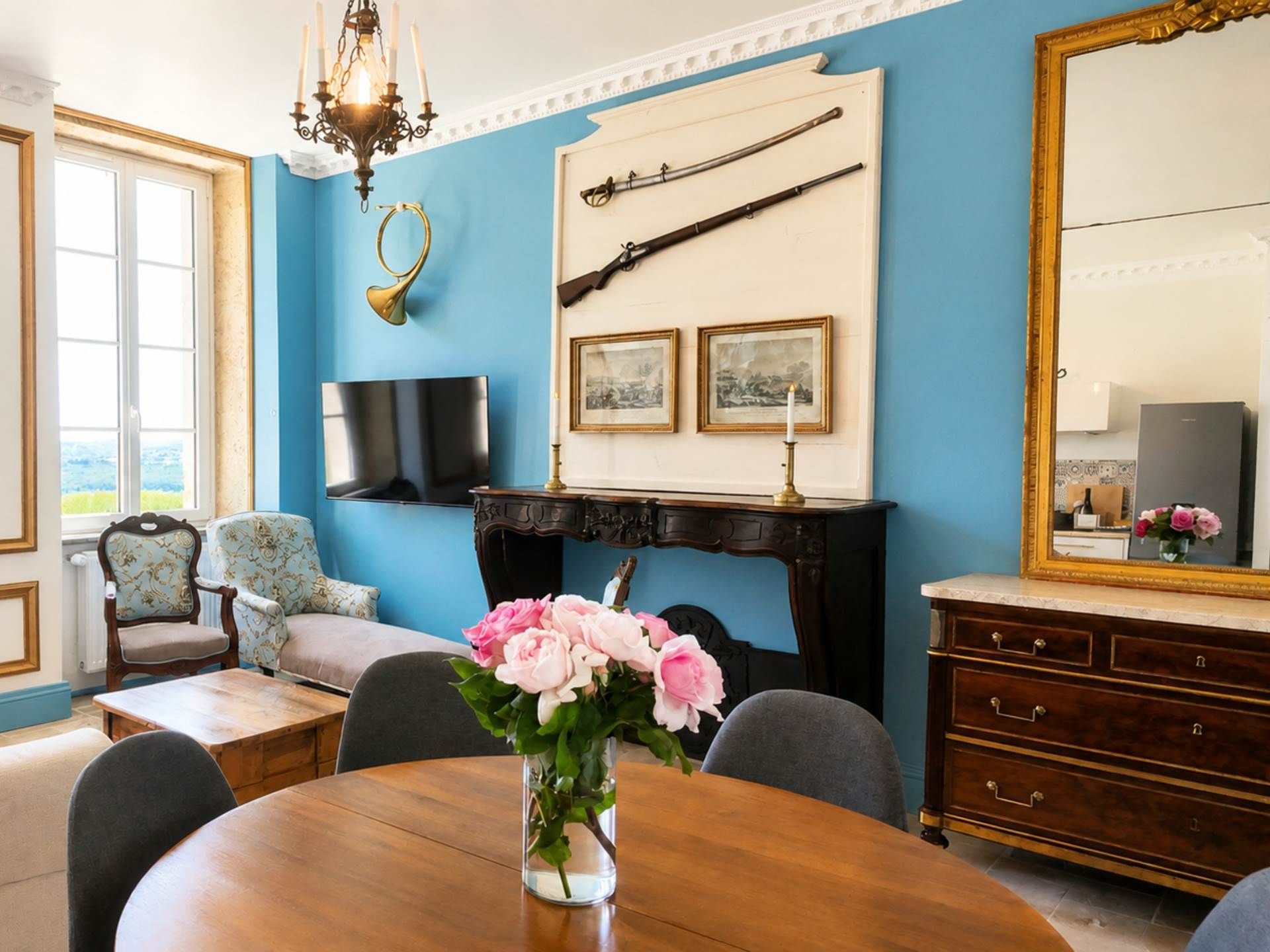 Une pièce avec un mur bleu, une cheminée, un fusil accroché au mur, un téléviseur, une table à manger ornée d’un vase de fleurs et un grand miroir.