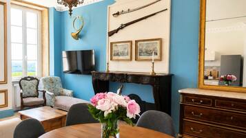 Une pièce avec un mur bleu, une cheminée, un fusil accroché au mur, un téléviseur, une table à manger ornée d’un vase de fleurs et un grand miroir.