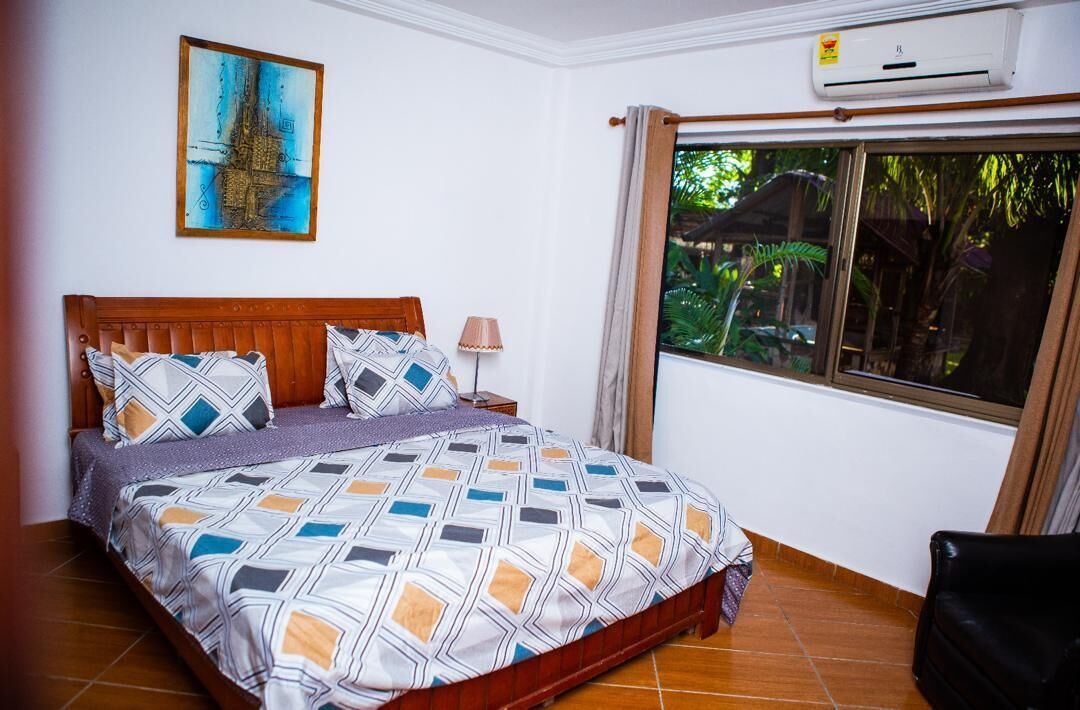 Um quarto com cama, mesa de cabeceira, abajur, janela com cortinas e ar-condicionado fixado na parede.