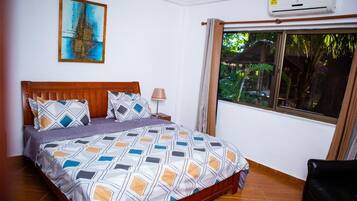 Um quarto com cama, mesa de cabeceira, abajur, janela com cortinas e ar-condicionado fixado na parede.