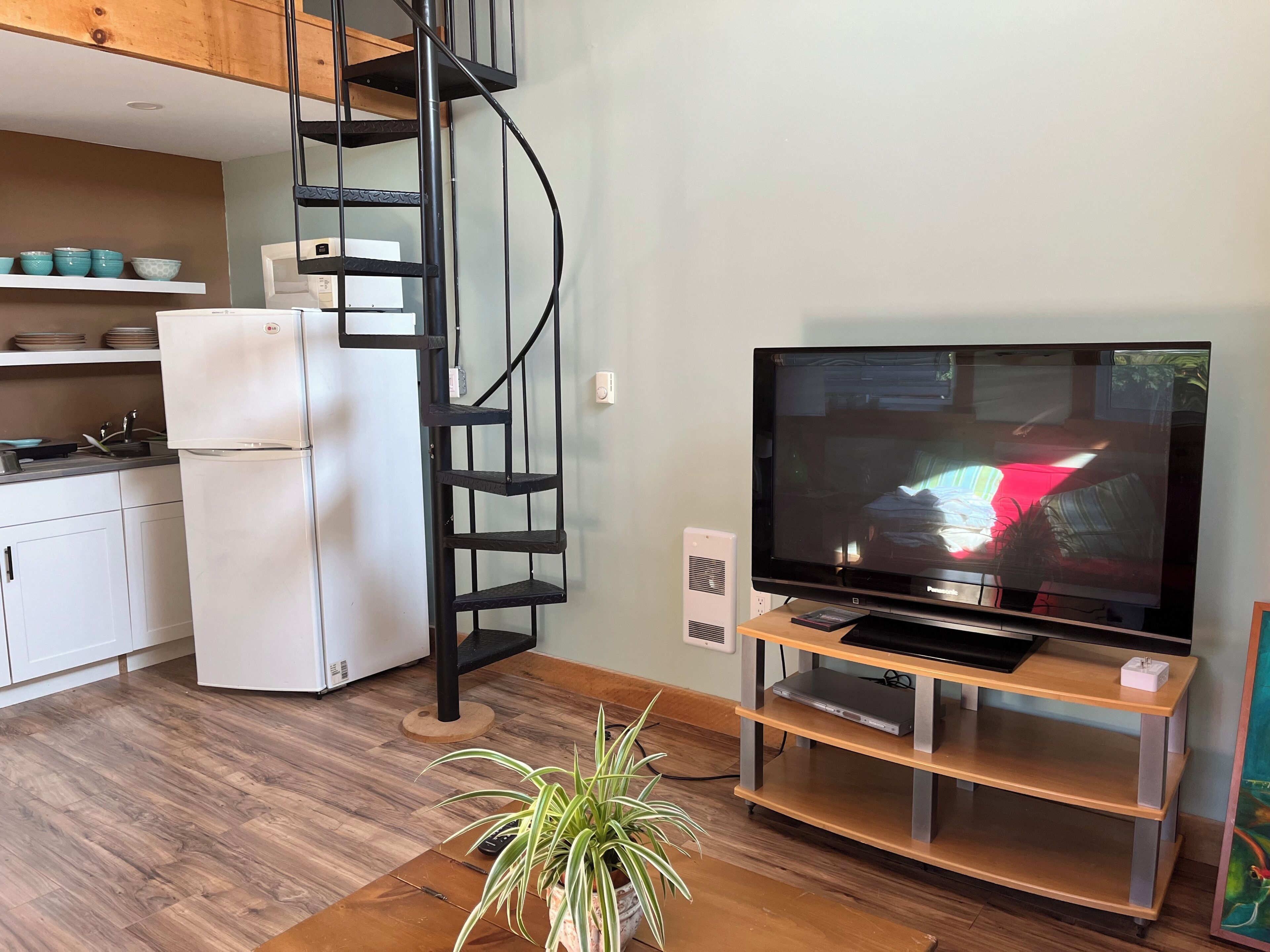 A modern living room with a flat-screen TV, a wooden entertainment unit, a spiral staircase, and a kitchen area with white cabinets and a refrigerator.