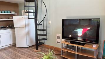 A modern living room with a flat-screen TV, a wooden entertainment unit, a spiral staircase, and a kitchen area with white cabinets and a refrigerator.
