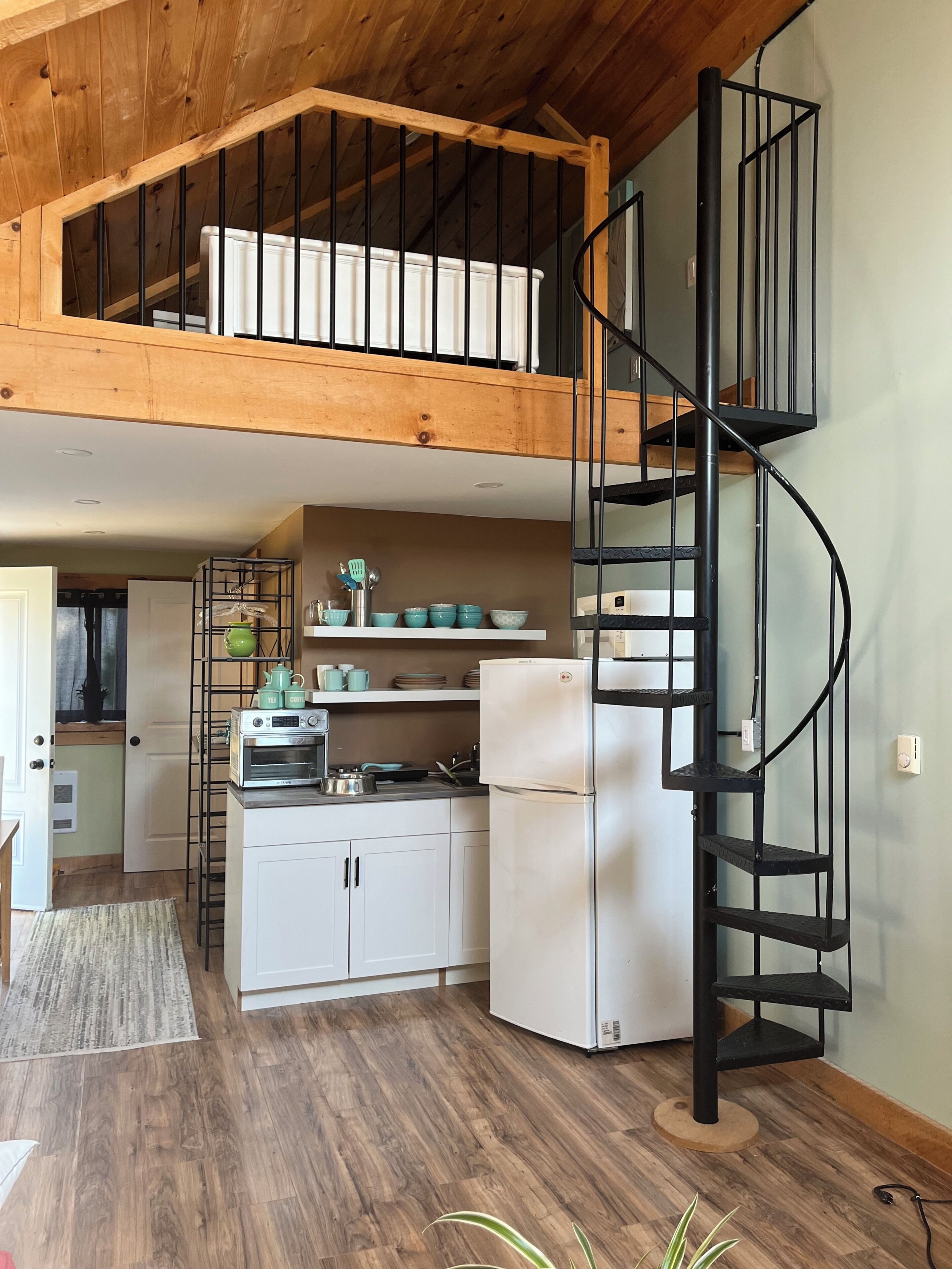 A kitchen with a wooden ceiling, a black spiral staircase, white cabinets, and a white refrigerator.