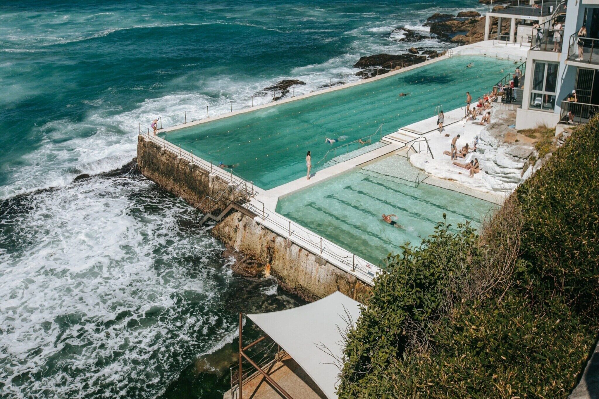 A large, rectangular swimming pool adjacent to a rocky coastline with waves crashing against the shore.