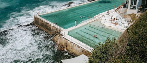 A large, rectangular swimming pool adjacent to a rocky coastline with waves crashing against the shore.