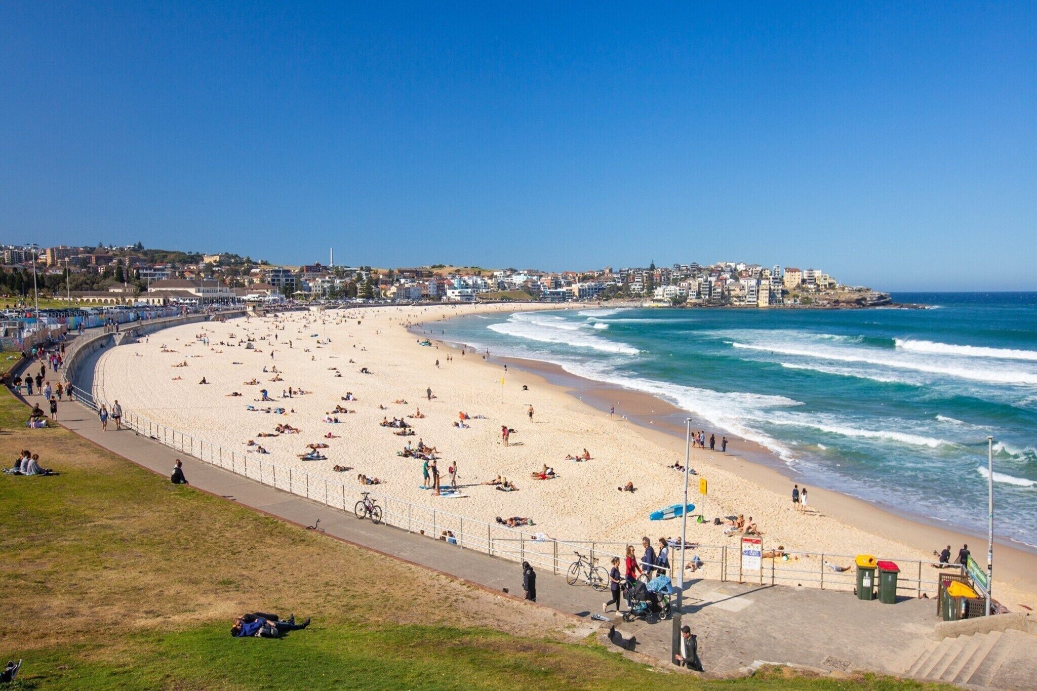 Una playa costera con un paseo marítimo, gente disfrutando en la playa y un paisaje urbano al fondo.