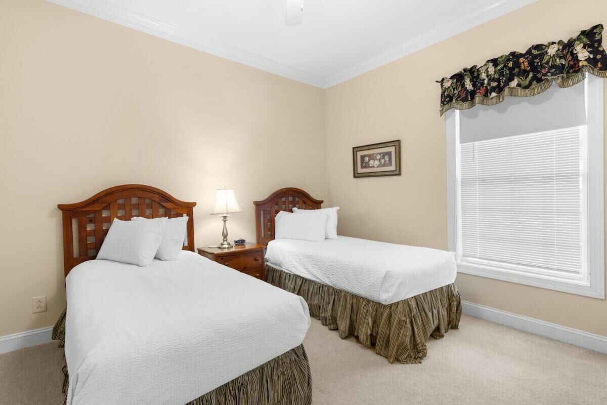 A bedroom with two beds, a nightstand with a lamp, a window with blinds, and a framed picture on the wall.