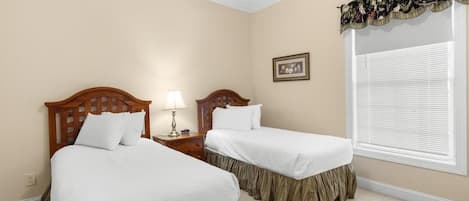 A bedroom with two beds, a nightstand with a lamp, a window with blinds, and a framed picture on the wall.