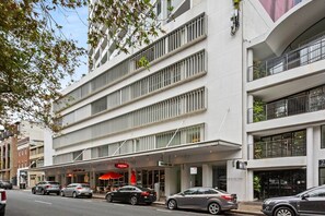 A multi-story hotel with a modern facade, a ground-level restaurant with a red awning, and several parked cars on the street. - Poolside Studio Retreat Near Oxford Street (Darlinghurst)