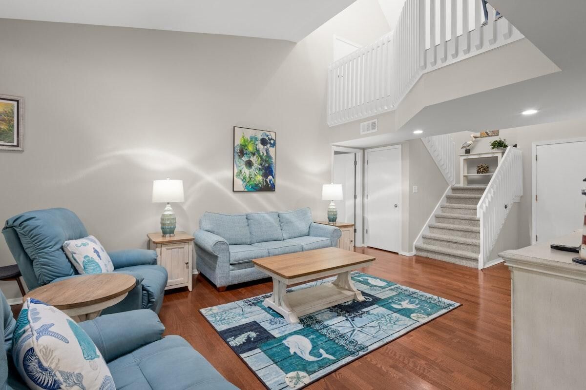 A spacious living room with a blue sofa set, a wooden coffee table, and a patterned rug. A staircase with a white railing leads to an upper level.