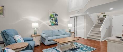 A spacious living room with a blue sofa set, a wooden coffee table, and a patterned rug. A staircase with a white railing leads to an upper level.