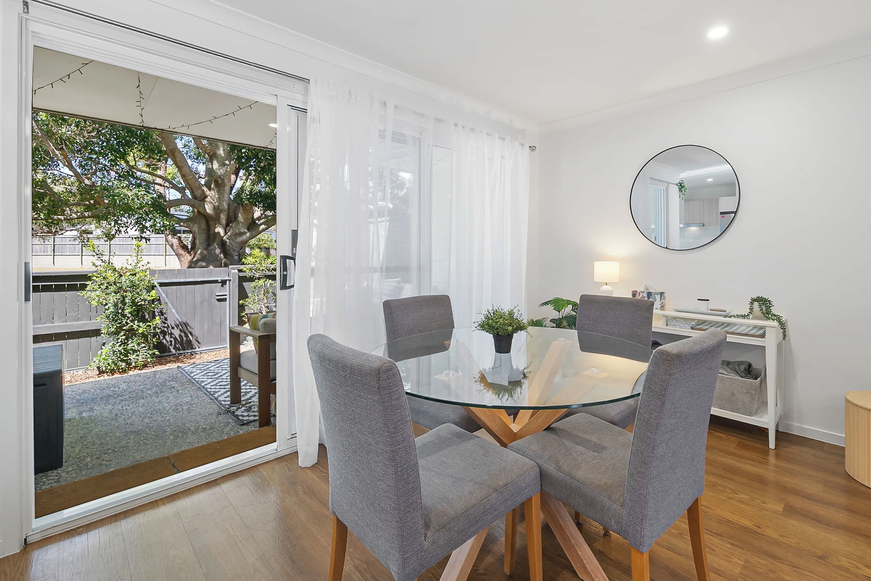 A modern dining area with a glass table and grey upholstered chairs, a round mirror on the wall, and a view of a tree outside.