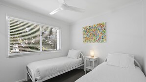 Una habitación con dos camas, un ventilador de techo, una ventana con persianas, una mesita de noche con lámpara y un cuadro abstracto colorido en la pared.