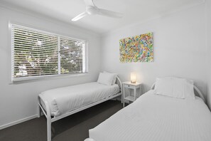A room with two beds, a ceiling fan, a window with blinds, a bedside table with a lamp, and a colorful abstract painting on the wall.