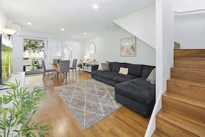 A modern living room with a sofa, a dining table with chairs, a staircase, and a view of the outdoors.