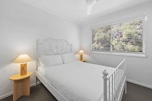 A bedroom with a white bed, two bedside lamps, a window with blinds, and a white ceiling fan.
