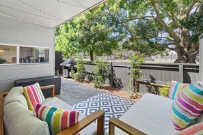 A covered outdoor seating area with a patterned rug, a barbecue grill, and a view of a backyard with trees and a fence.