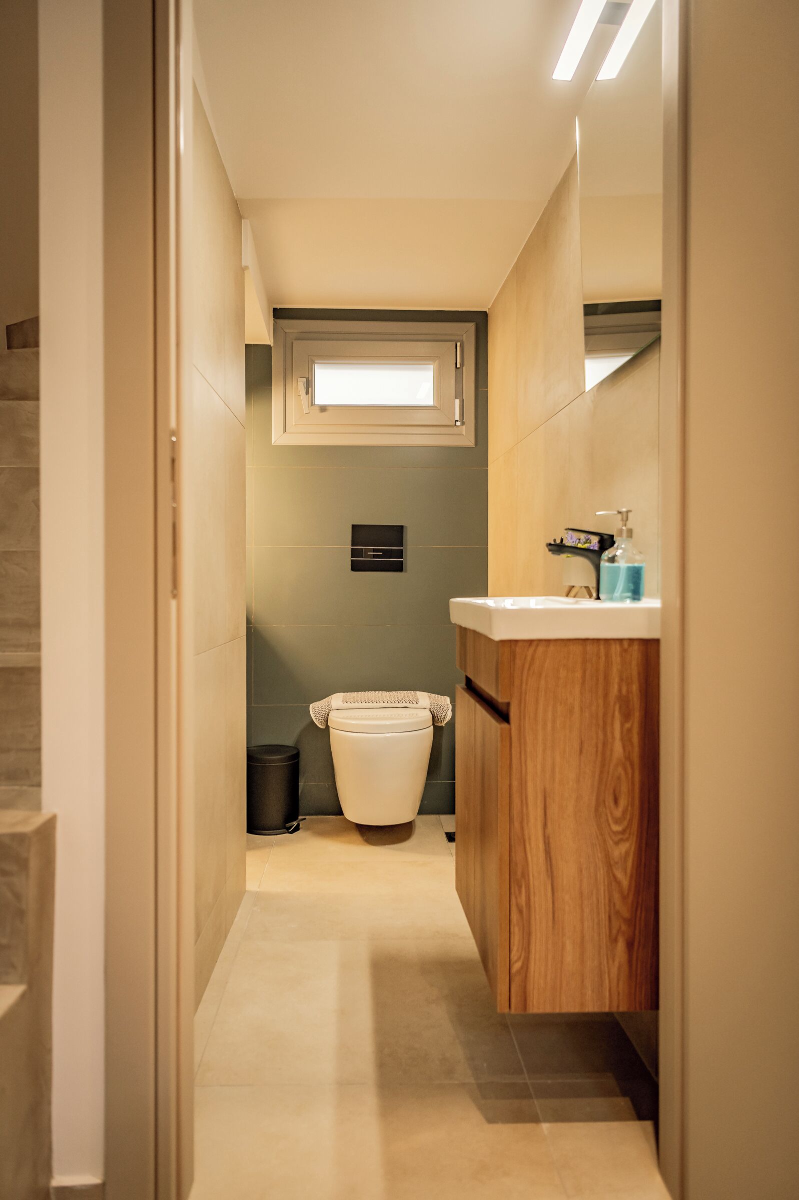 A narrow hallway leading to a bathroom with a toilet, a trash bin, and a wooden vanity with a mirror.