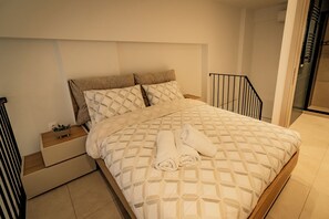 Ein ordentlich bezogenes Bett mit einem geometrisch gemusterten Überwurf, ein Paar zusammengerollte Handtücher und ein Nachttisch mit einer kleinen Pflanze.