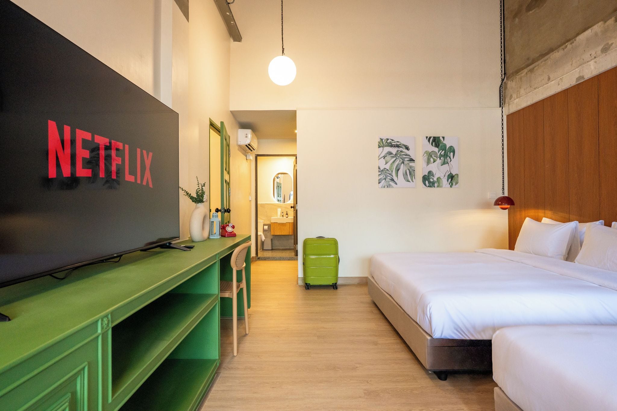 A hotel room with a bed, a desk, a green suitcase, a wall-mounted TV displaying 'NETFLIX', and a bathroom visible through an open door.