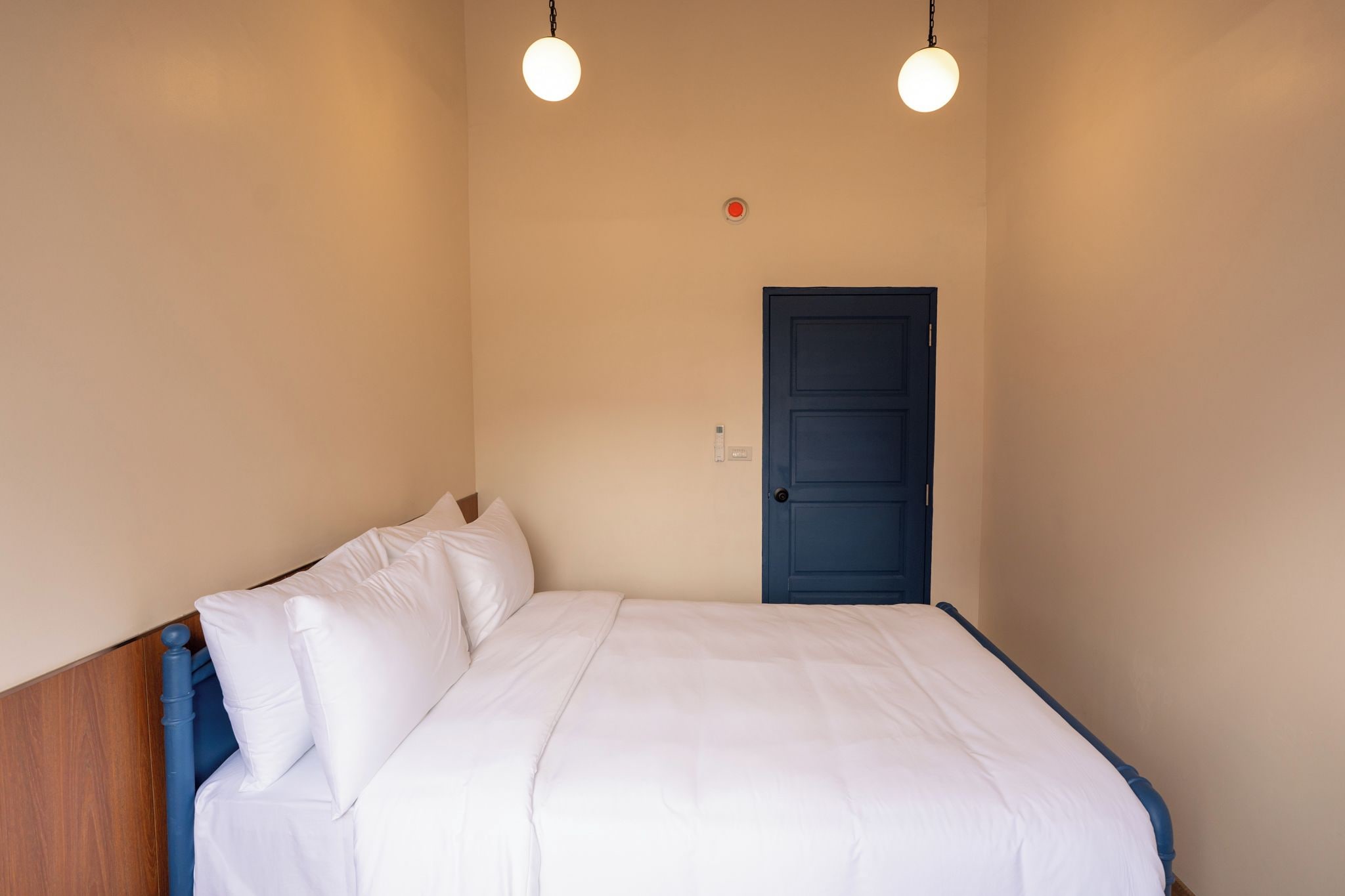 A single bed with white bedding in a room with beige walls, a blue door, and two hanging light fixtures.