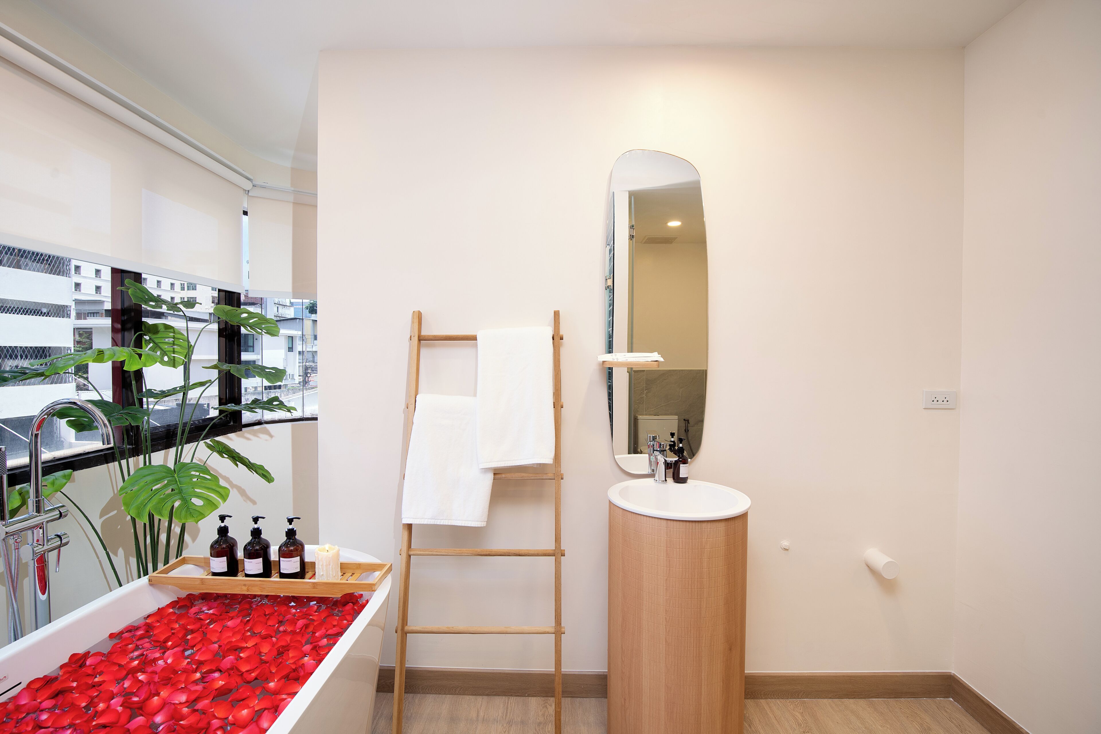 Un baño con una bañera llena de pétalos de rosa, una escalera de madera, un lavabo con espejo y una ventana con vistas a edificios.