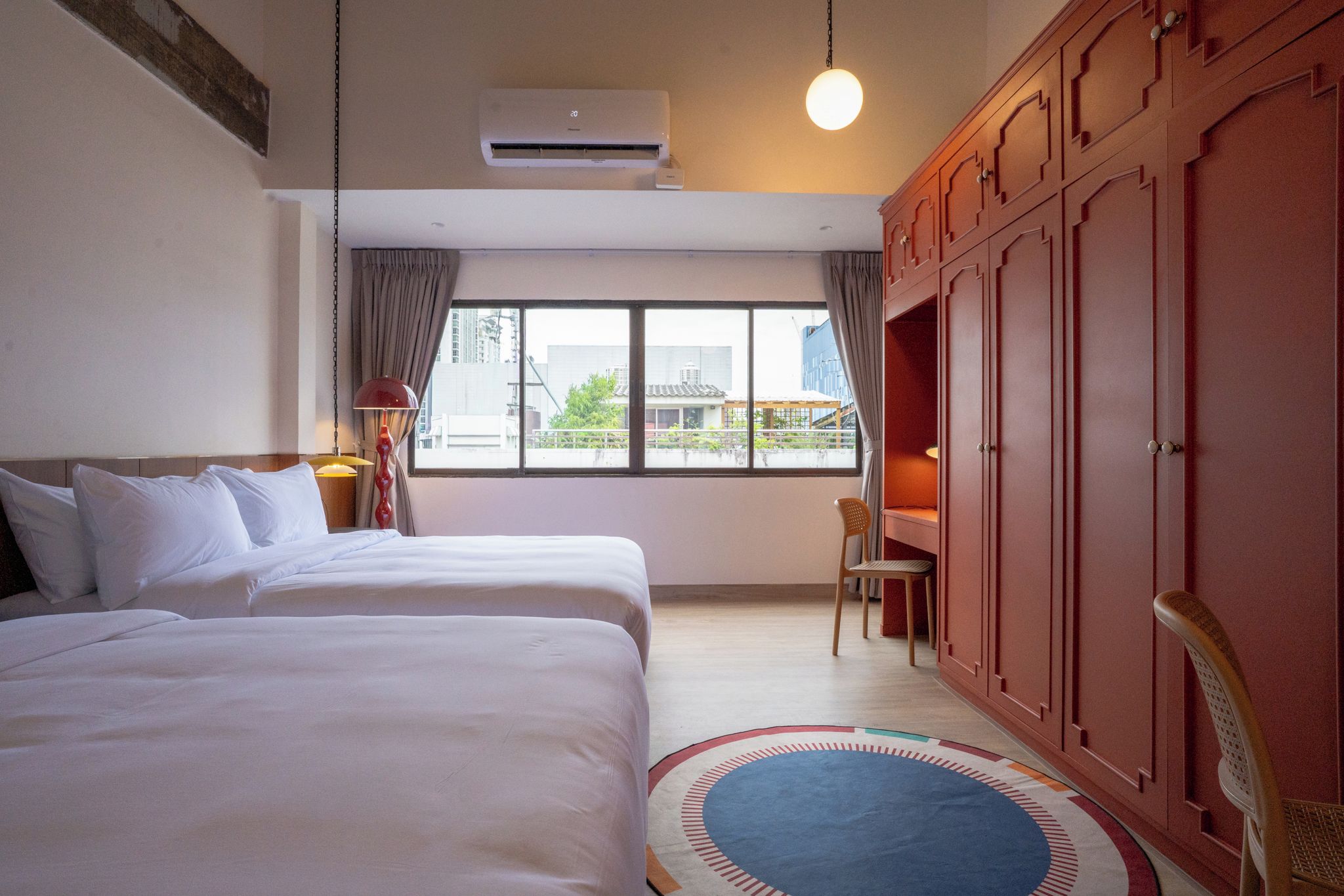 A hotel room with two beds, a desk, a chair, an air conditioning unit, and a view of buildings outside.