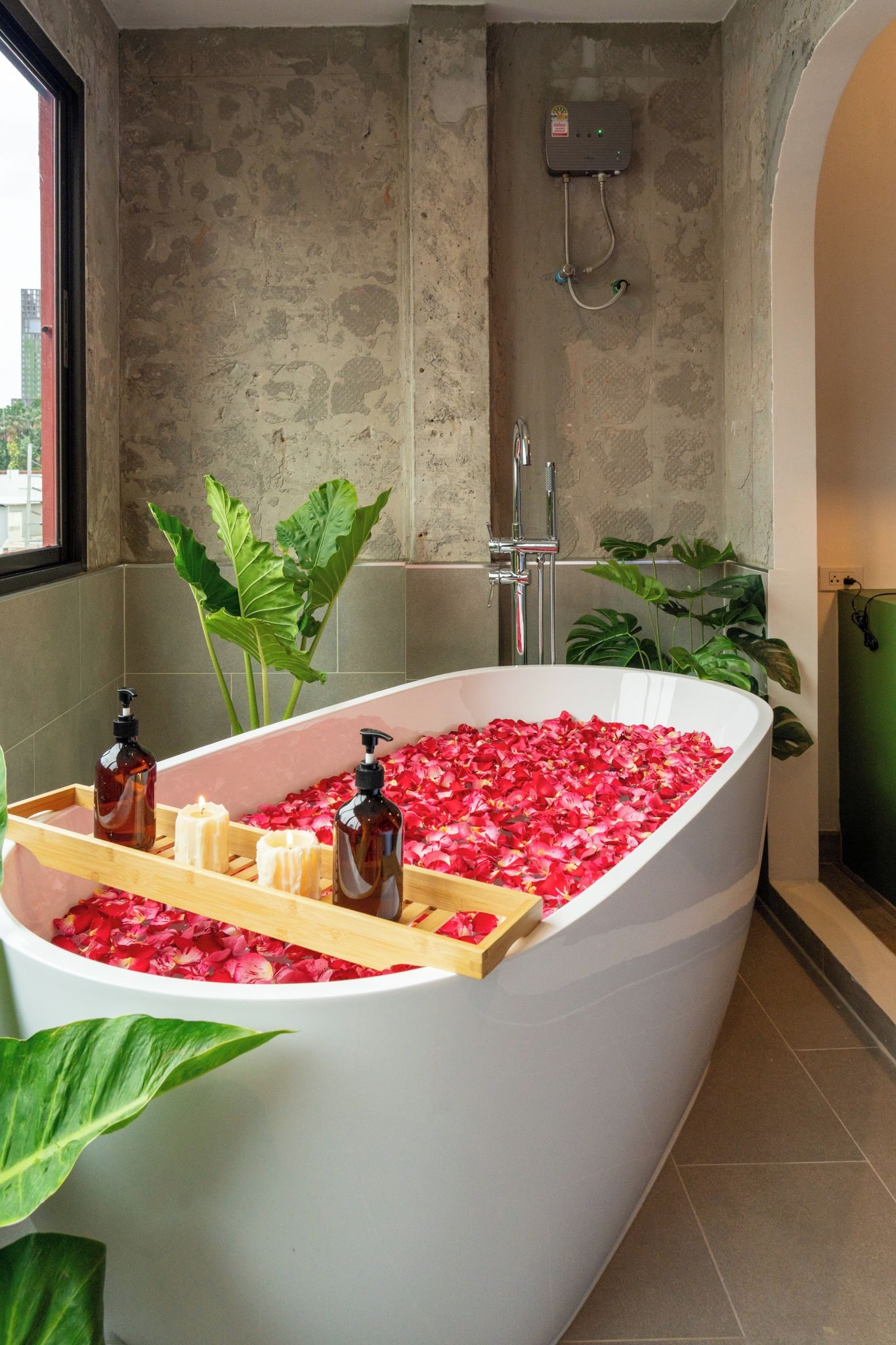 Eine Badewanne, gefüllt mit Rosenblättern und Badesachen, mit einem Fenster und Pflanzen im Hintergrund.