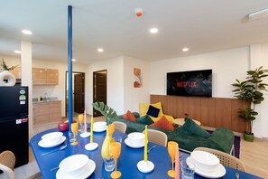 Un salon moderne avec un coin repas, un téléviseur à écran plat fixé au mur et une cuisine visible au fond.