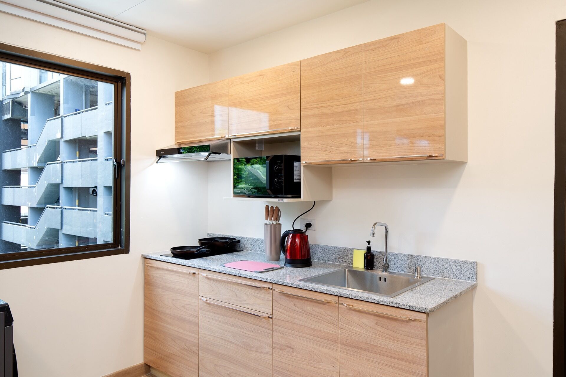 A compact kitchen with wooden cabinets, a built-in microwave, a stainless steel sink, and a window overlooking a parking structure.